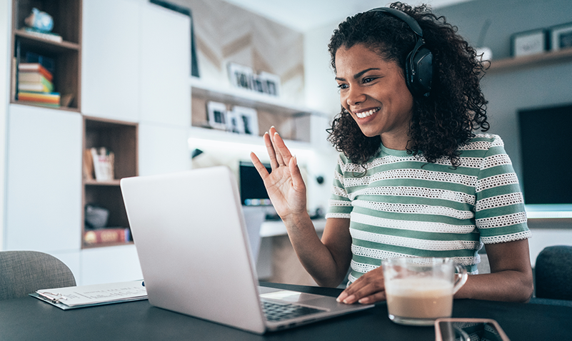 A female employee working from home wears headphones as she smiles and waves to her laptop camera.