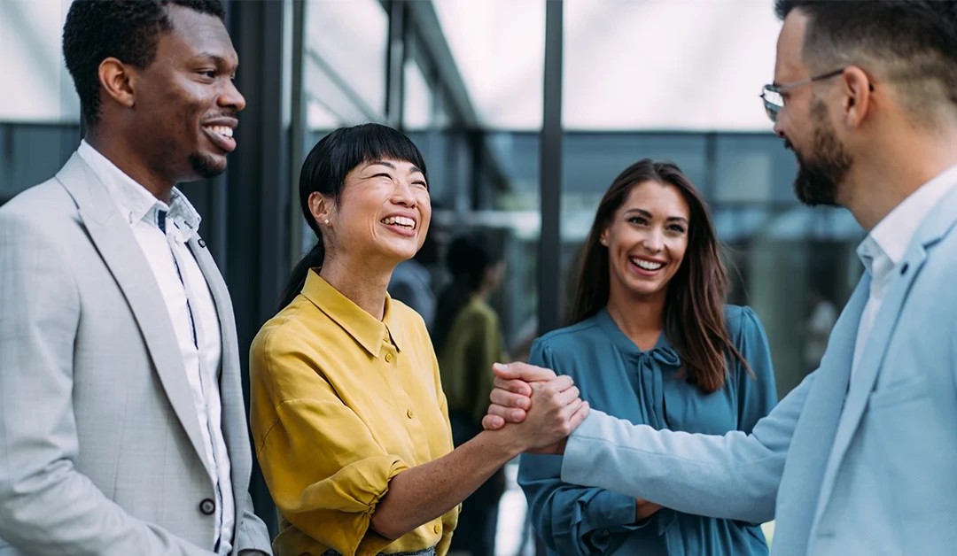 four professionals talking and smiling. A woman in a yellow shirt shaking hands with a man in a grey suit.